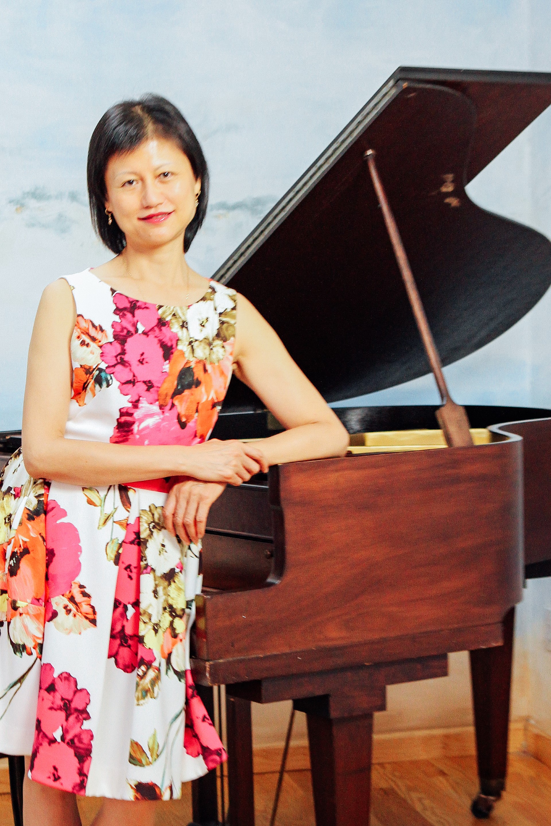 Woman leaning against piano with a floral dress and gray background.