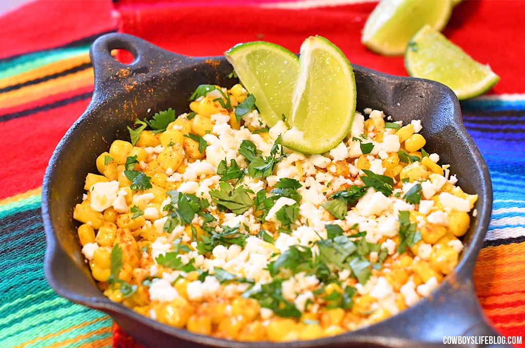 Mexican street corn topped with cilantro, cotija cheese and lime wedges in an iron skillet