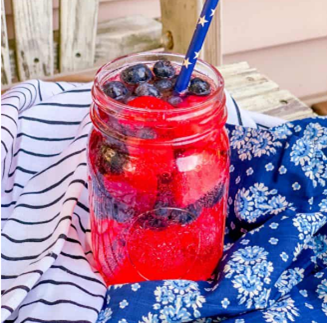 Picture of a mason jar atop a table with artfully strewn summer blue table covers. The mason jar contains an ambrosia type drink topped with berries.