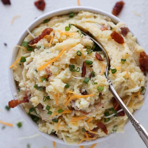 a white bowl filled with mashed potatoes topped with cheese, bacon and chives with a large silver spoon in the middle of the bowl