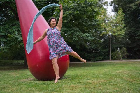 Woman dancing on green grass with green trees in the background with a scarf and a red sculpture in the background also.