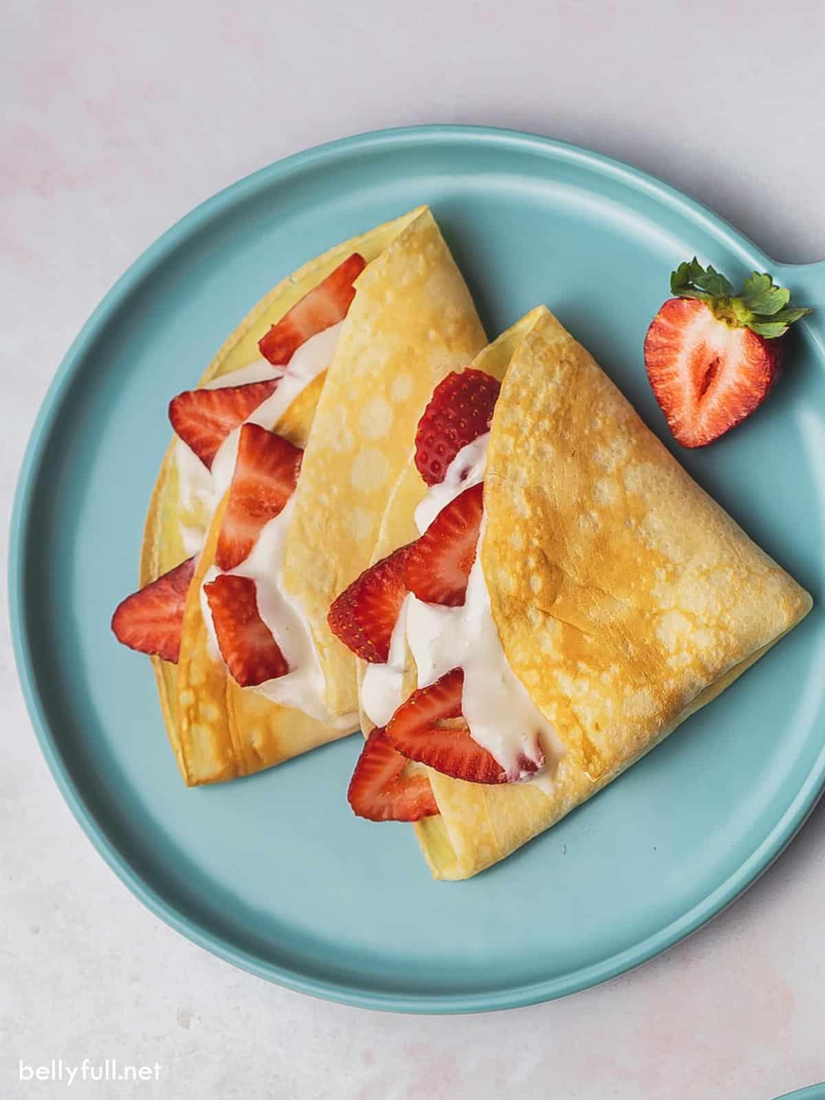 Twin medley of strawberries swaddled in cream and crepes rest upon an ice blue plate. 