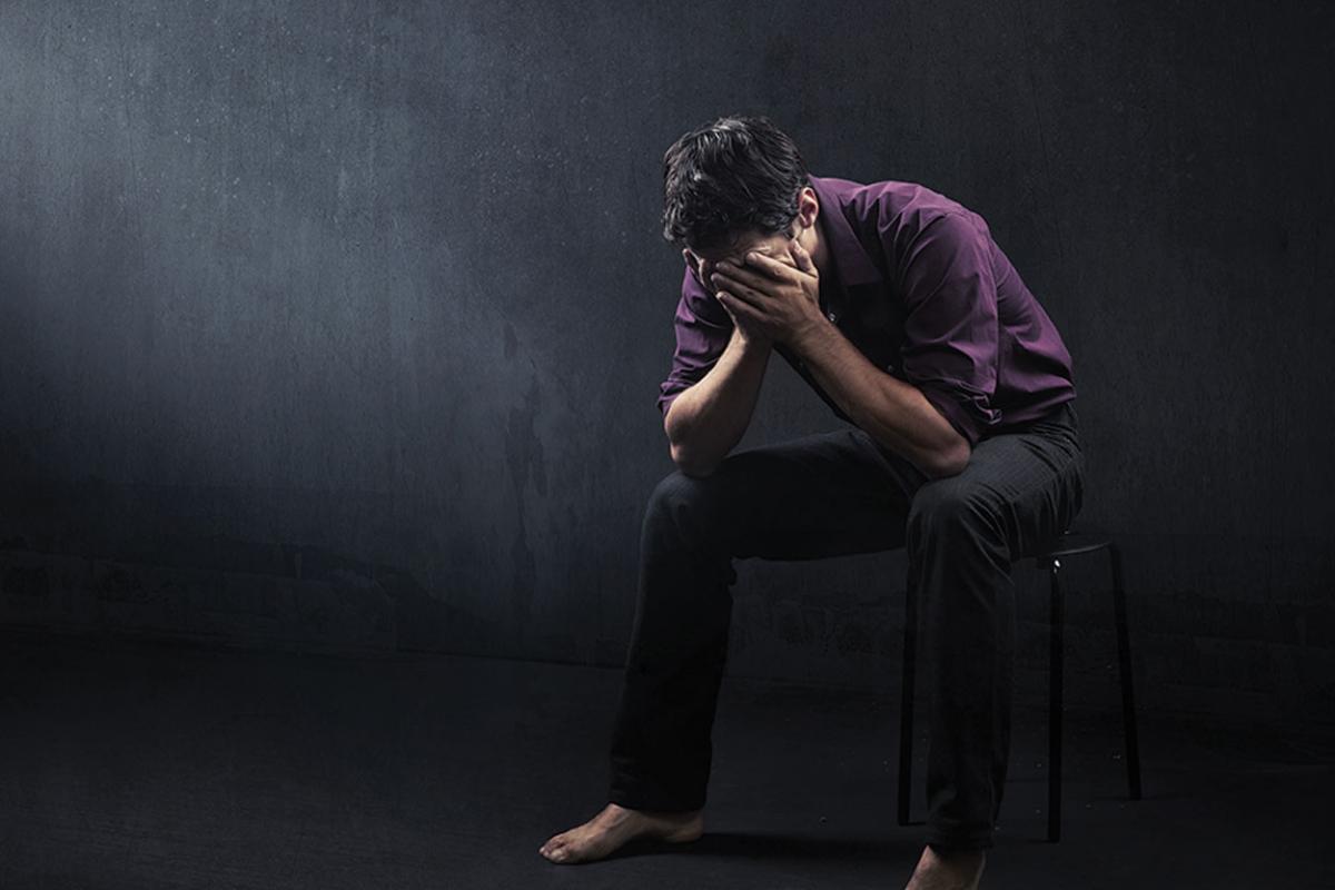 A man covering his face upset and black background and purple shirt.