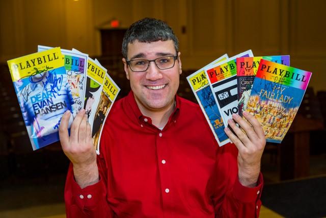 A man in a red shirt with glasses holding Broadway Play Bills in both hands.