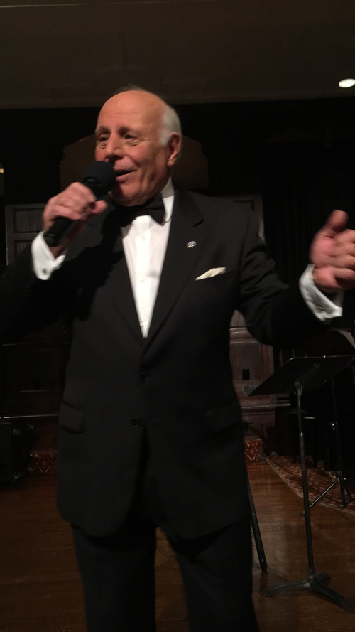 Man in a black tuxedo singing with a black background.