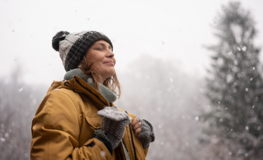 A woman smiling standing in the snow.  White and gray background with trees.