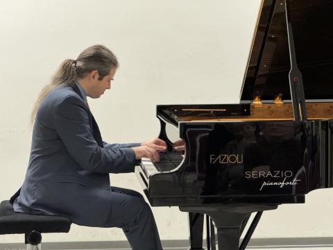 Itay Goren sitting at piano playing in a black suit with a white background.