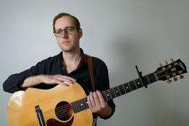 Man in glasses holding a guitar with a white wall background.