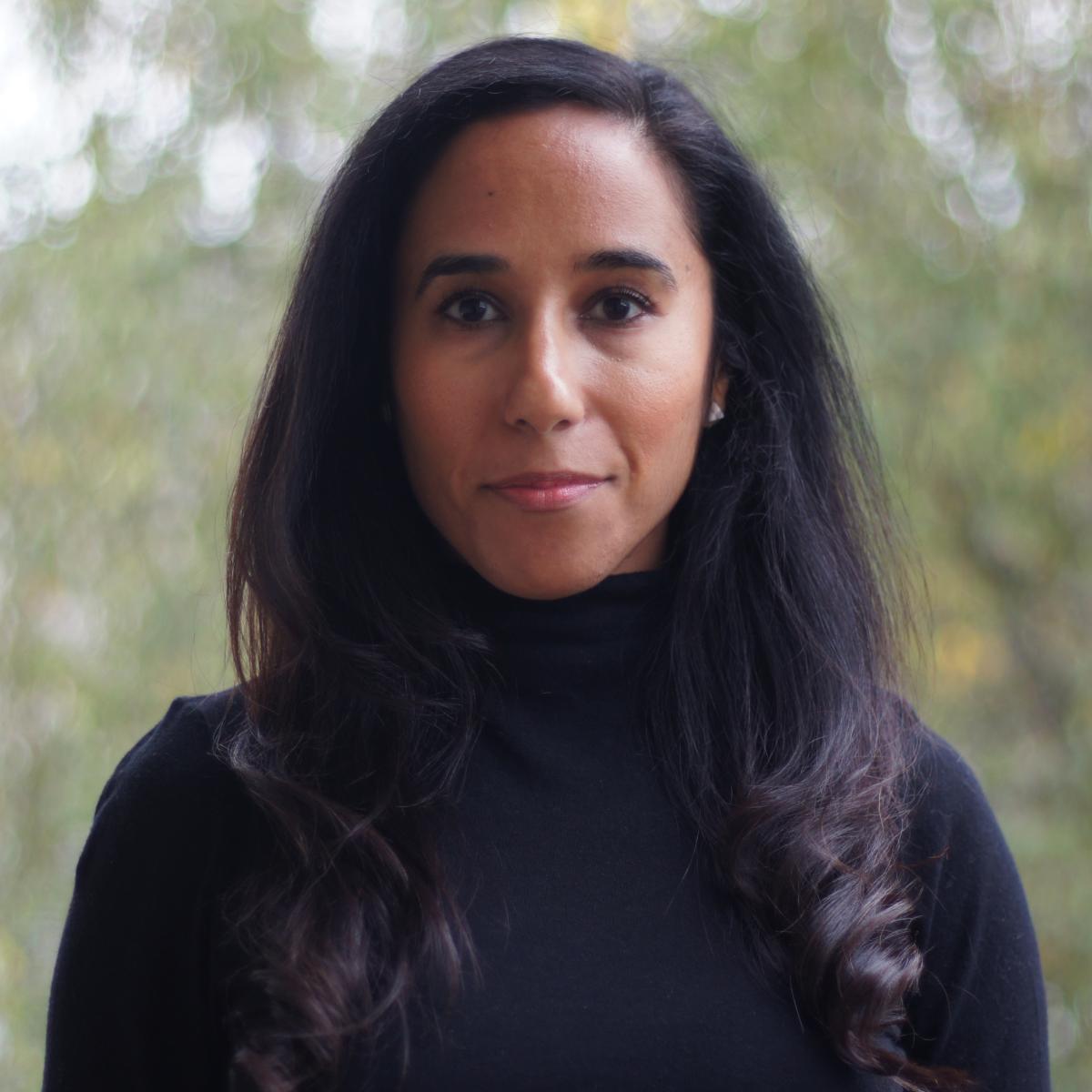 A woman with long black hair in a black turtle neck sweater with green trees in the background.