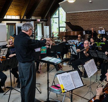 Conductor wearing black in front of musicians with music books on stands.